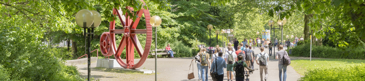 Studierende laufen über den Campus Nord. Es ist eine große rote Skulptur in Form von großen Zahnrädern zu erkennen.