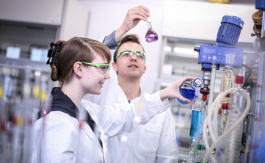 Zwei Auszubildende in einem chemischen Labor mit weißen Kitteln, einer gießt blaue Flüssigkeit in ein Destilliergerät.