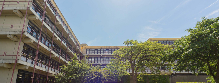 Chemiegebäude: Ein großes, breites, graues Gebäude mit Fensterfronten und Balkonen vor den Fenstern, die mit roten Metallstangen abgegrenzt werden. In der Mitte eine Treppe, auf der Menschen sitzen und links vor der Treppe Fahrräder in Fahrradständern.