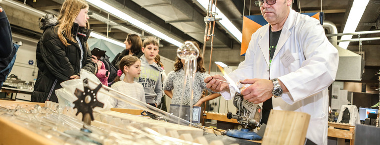A man in a white lab coat stands at a table in the lab and children stand around the table.