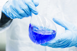 Two hands wearing blue gloves are holding a volumetric flask with a dark blue liquid inside.