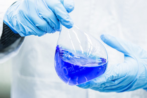 Zwei Hände in blauen Gummihandschuhen halten einen Messkolben, der mit blauer Flüssigkeit gefüllt ist.
